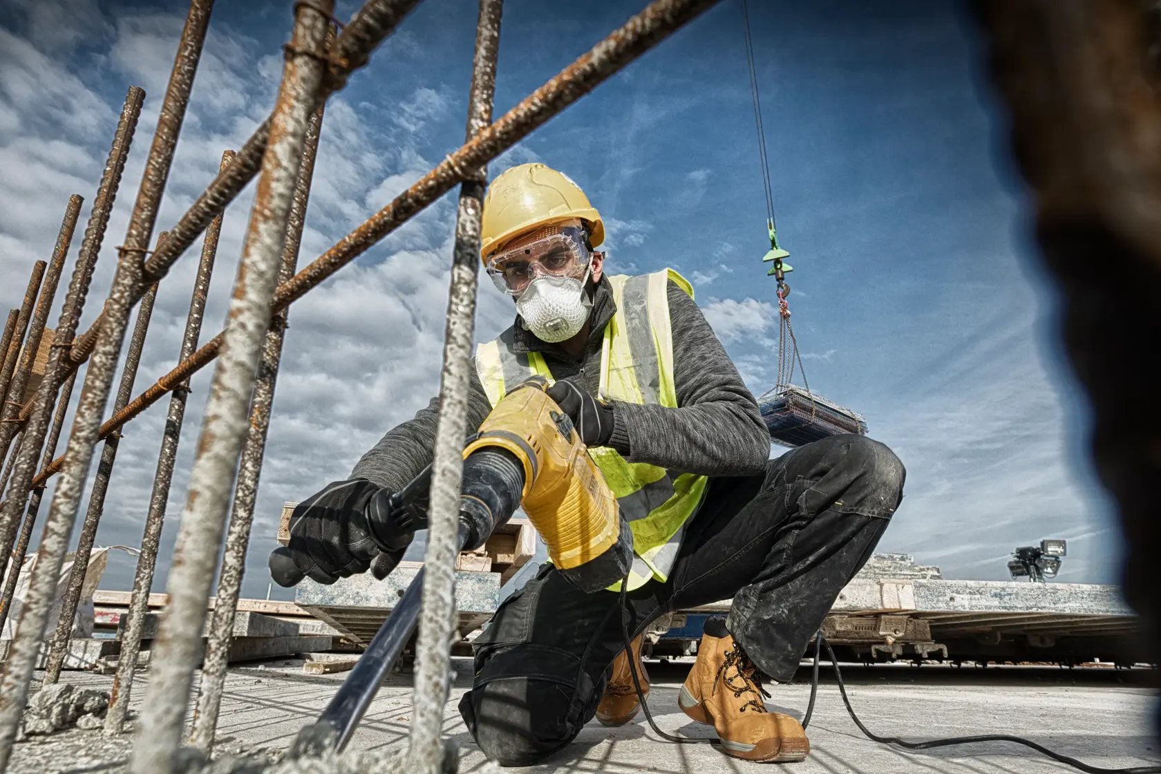 Worker uses a DEWALT drill on a ceiling at a construction site, wearing safety gear and a high-visibility vest.