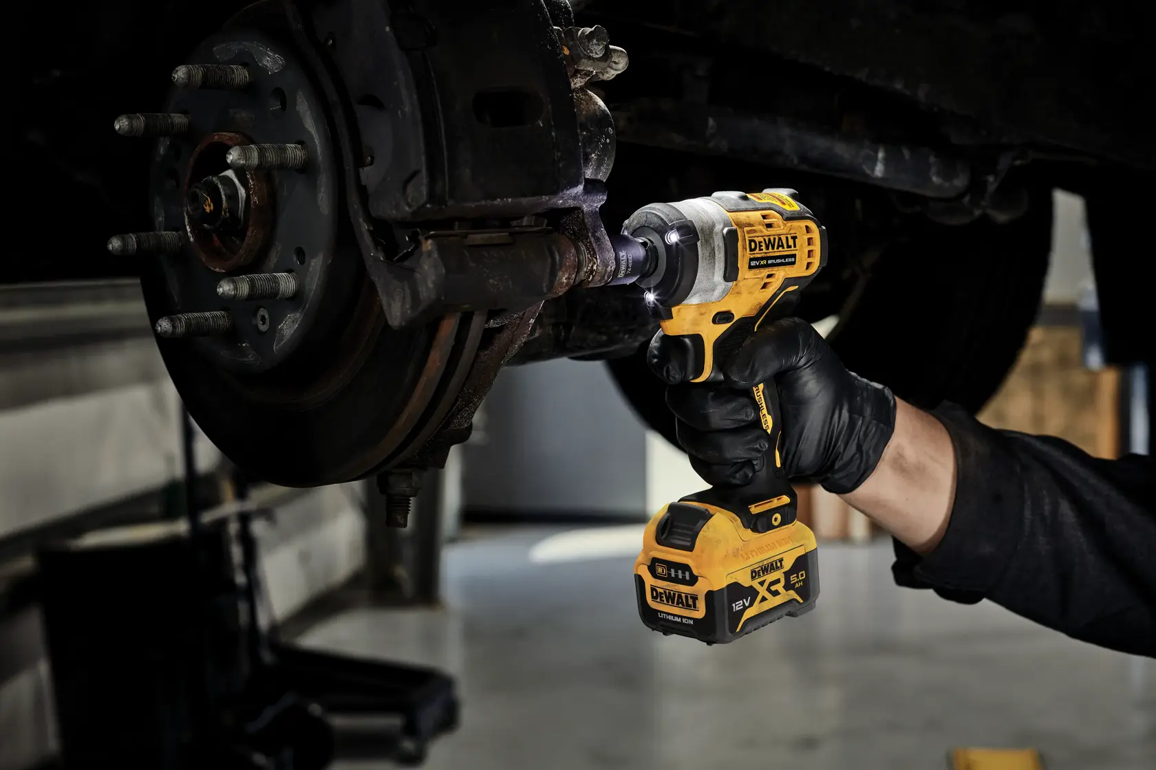Gloved hand using a DEWALT cordless impact wrench on a vehicle’s wheel hub in a garage.