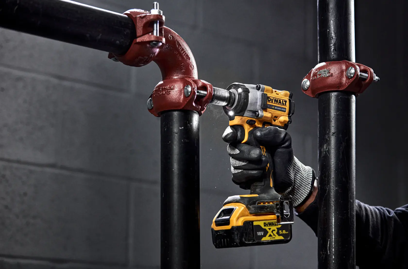 Eine Person mit Handschuhen verwendet einen kabellosen Schlagschrauber DEWALT DCF921NT, um eine Metallrohrverbindung in einer industriellen Umgebung festzuziehen oder zu lösen.