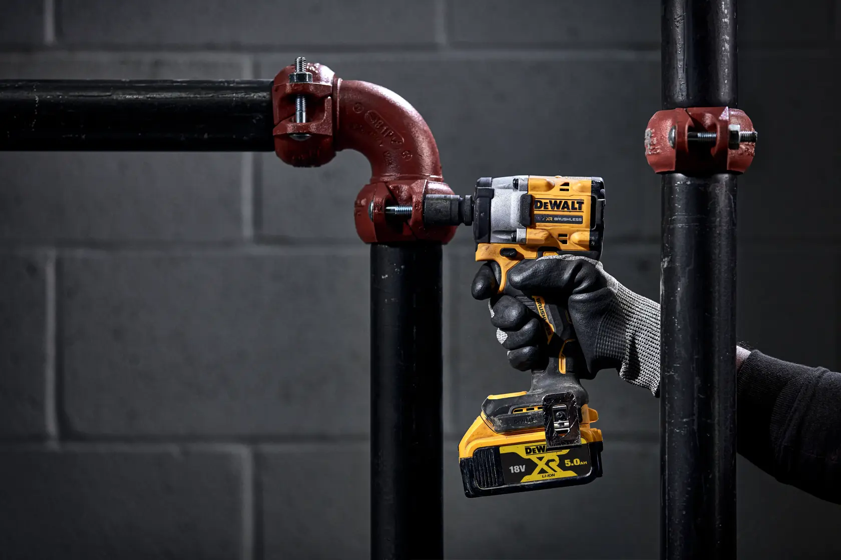 Una persona con guantes negros utiliza una llave de impacto inalámbrica DEWALT para apretar un perno en una unión de tubería metálica. La herramienta es amarilla y negra, y el paquete de batería es visible. El fondo muestra una pared de ladrillos grises y tuberías industriales.
