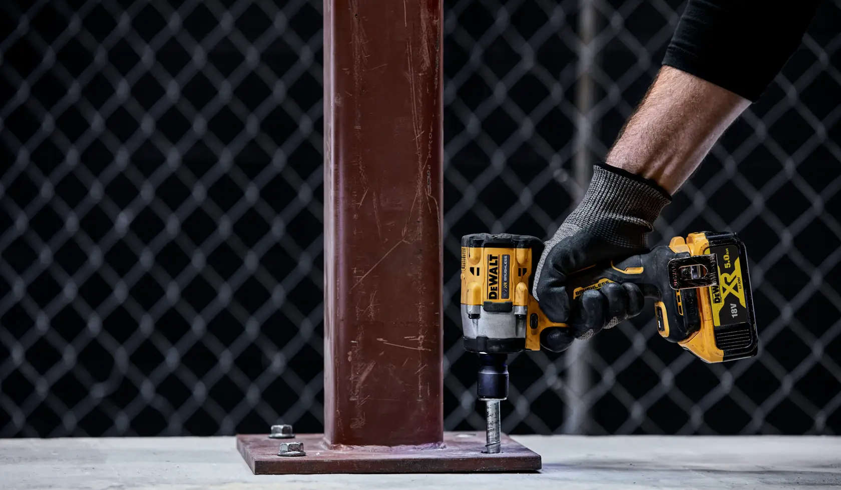 A person wearing a glove uses a DEWALT cordless impact wrench to tighten a bolt securing a metal beam to a concrete surface.