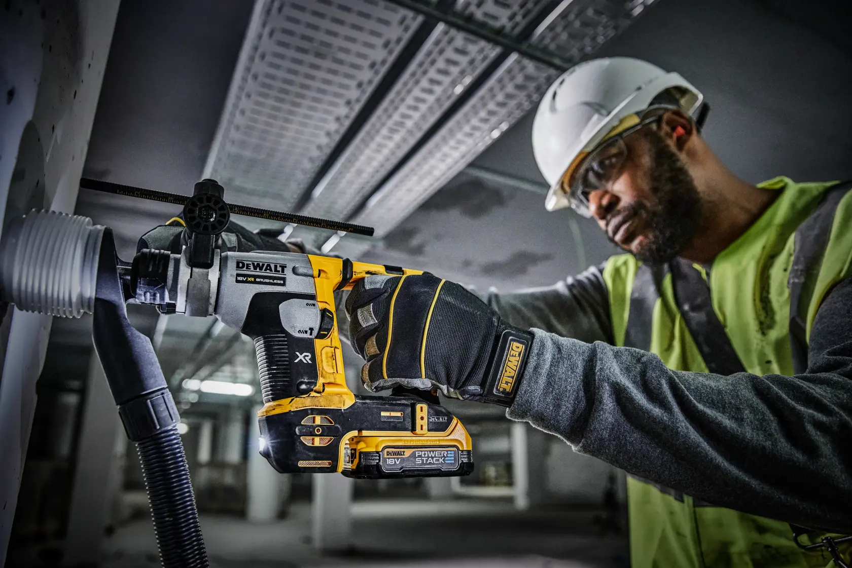 Una persona con casco de seguridad y chaleco de alta visibilidad está utilizando un taladro rotativo inalámbrico DEWALT para perforar una pared, con equipo de extracción de polvo acoplado. El taladro tiene las marcas XR y Power Stack.