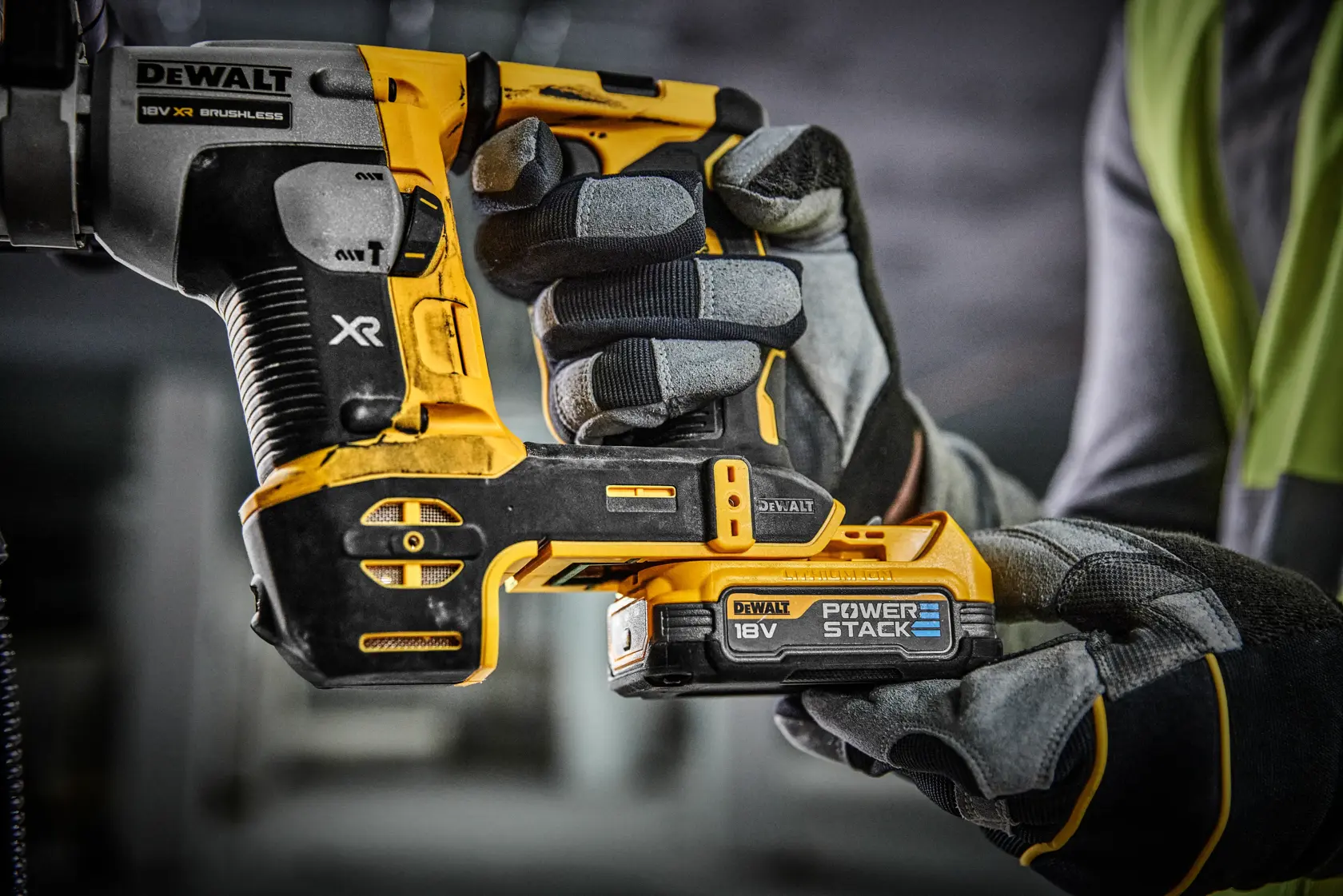 Primer plano de una persona con guantes de trabajo sosteniendo una herramienta eléctrica DEWALT 18V XR sin escobillas con una batería Power Stack en un entorno de taller o construcción.