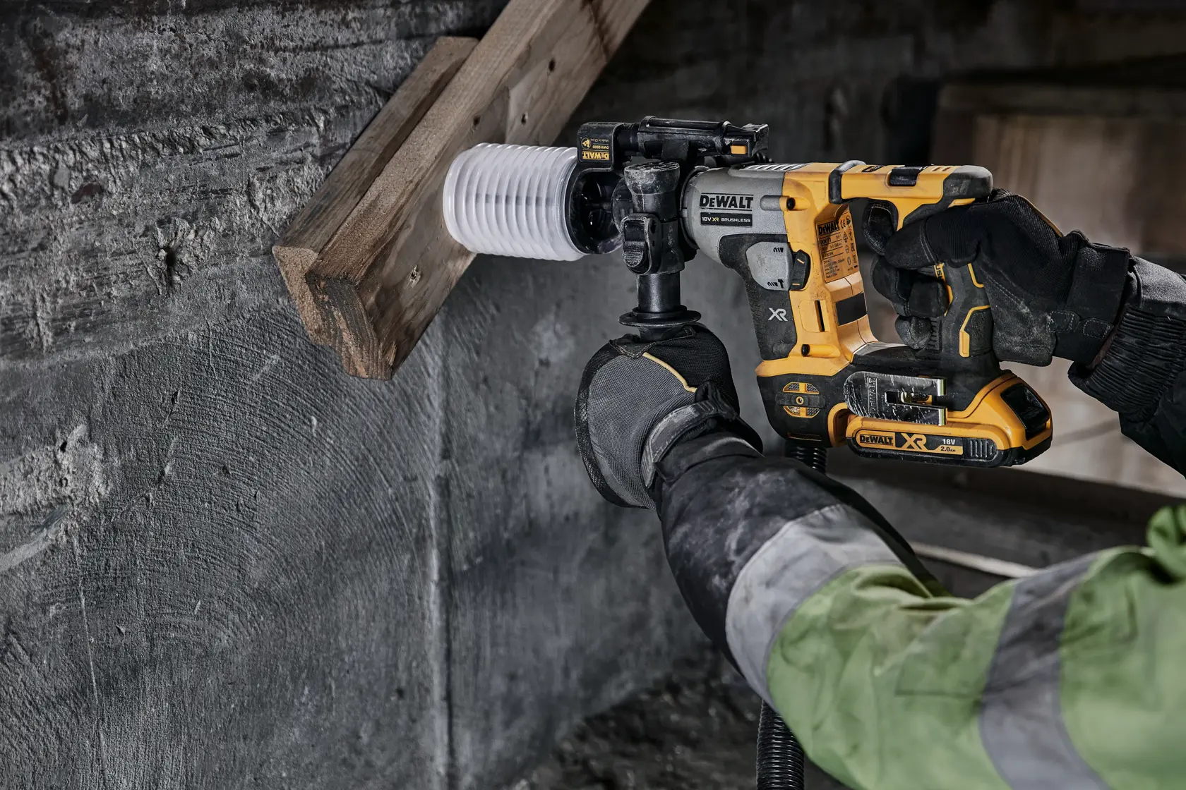 A DEWALT cordless hammer drill is used on concrete near wood, held by a gloved worker in a construction setting.