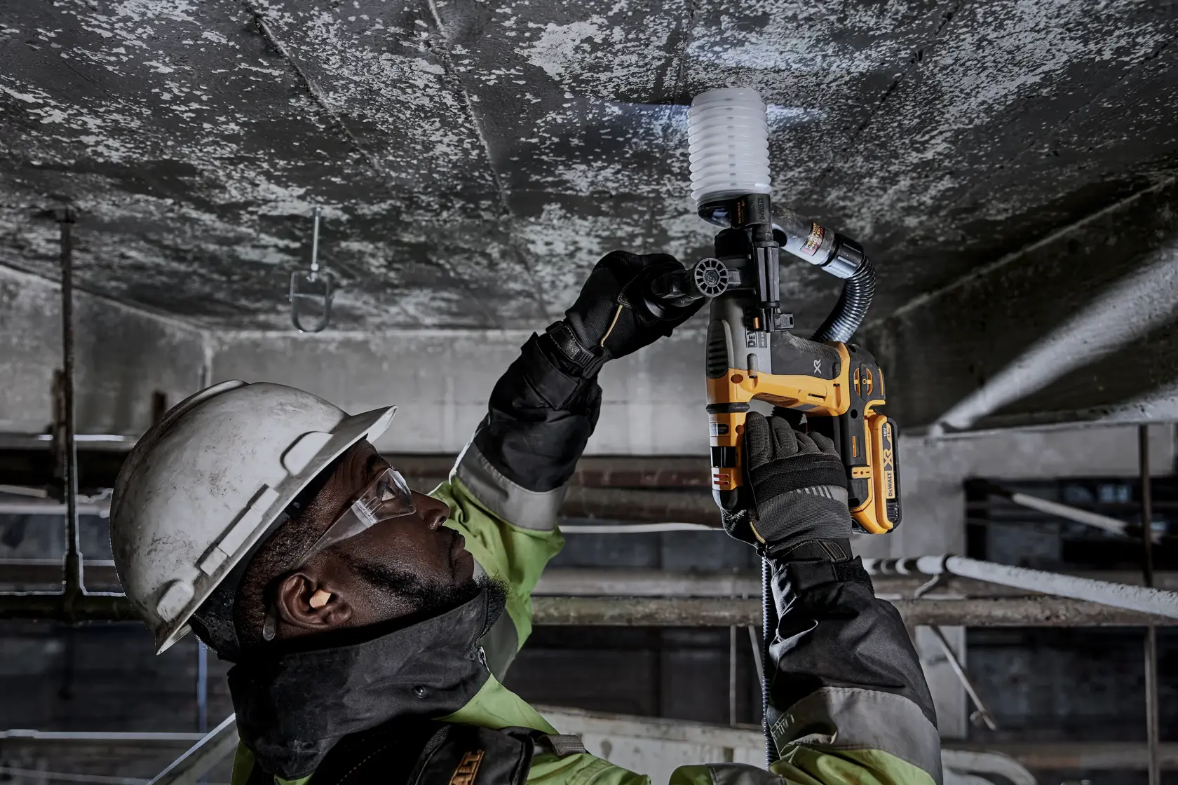 Person uses a DEWALT DCH172N rotary hammer drill on a ceiling, wearing safety gear and helmet at a construction site.