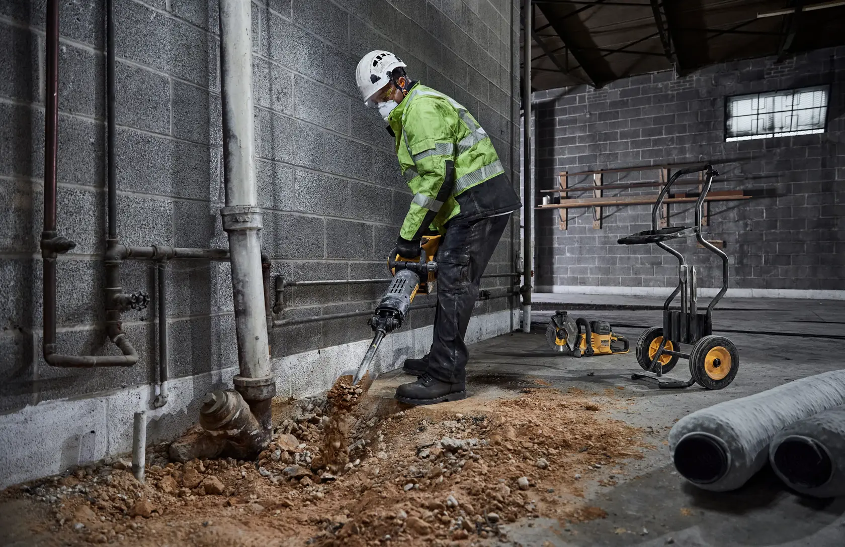 Una persona con equipo de seguridad utiliza un martillo demoledor DEWALT para romper un suelo de hormigón cerca de una pared en un entorno industrial. Herramientas y equipos de construcción son visibles al fondo.