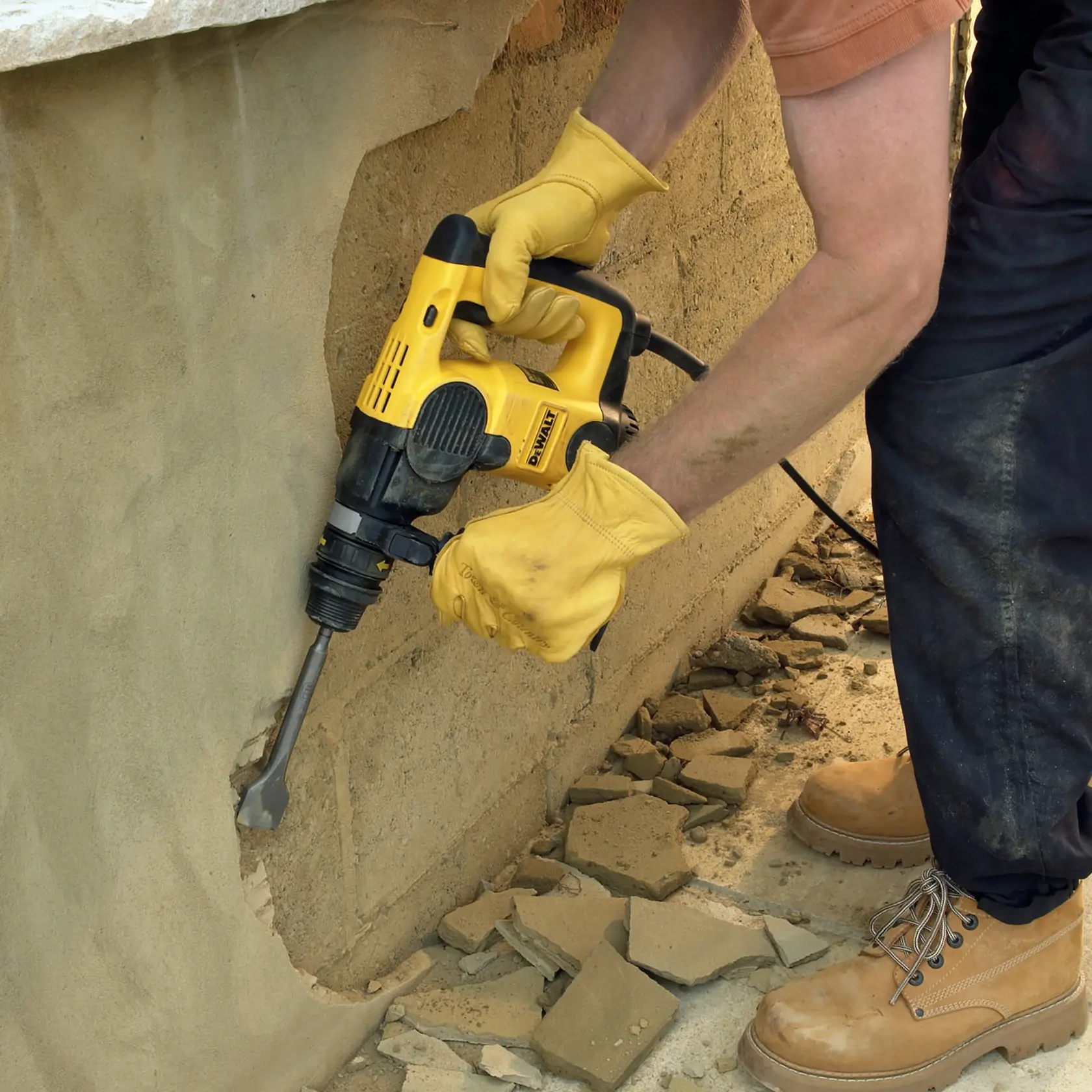 Person uses DEWALT hammer drill to break plaster wall, wearing yellow gloves and boots amid rubble.