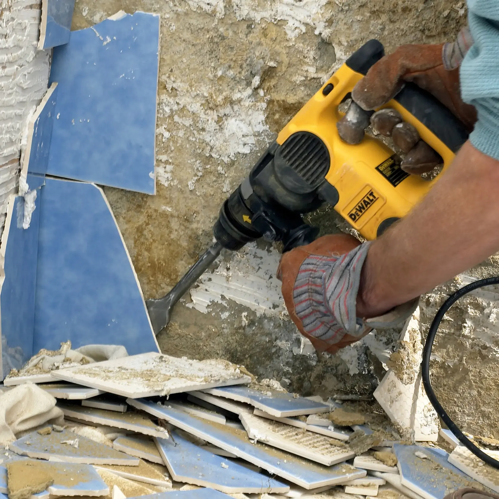 Worker uses a DEWALT tool to remove blue wall tiles, breaking them off a rough surface.