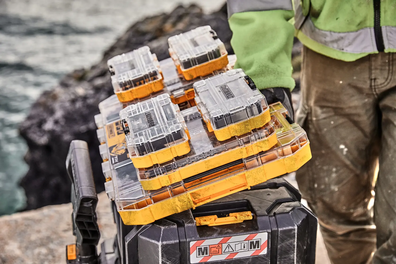 DEWALT DT70762 stackable tool cases with transparent lids, held by a worker near water and rocks.