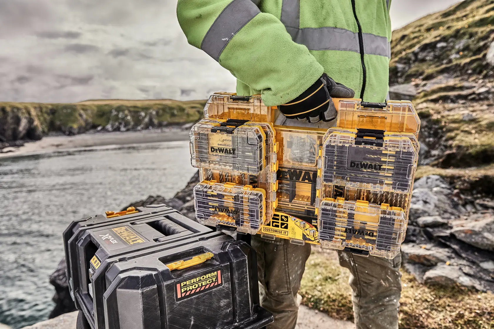 Person holding DEWALT DT70762 tool storage case next to another DEWALT toolbox outdoors near rocky water.
