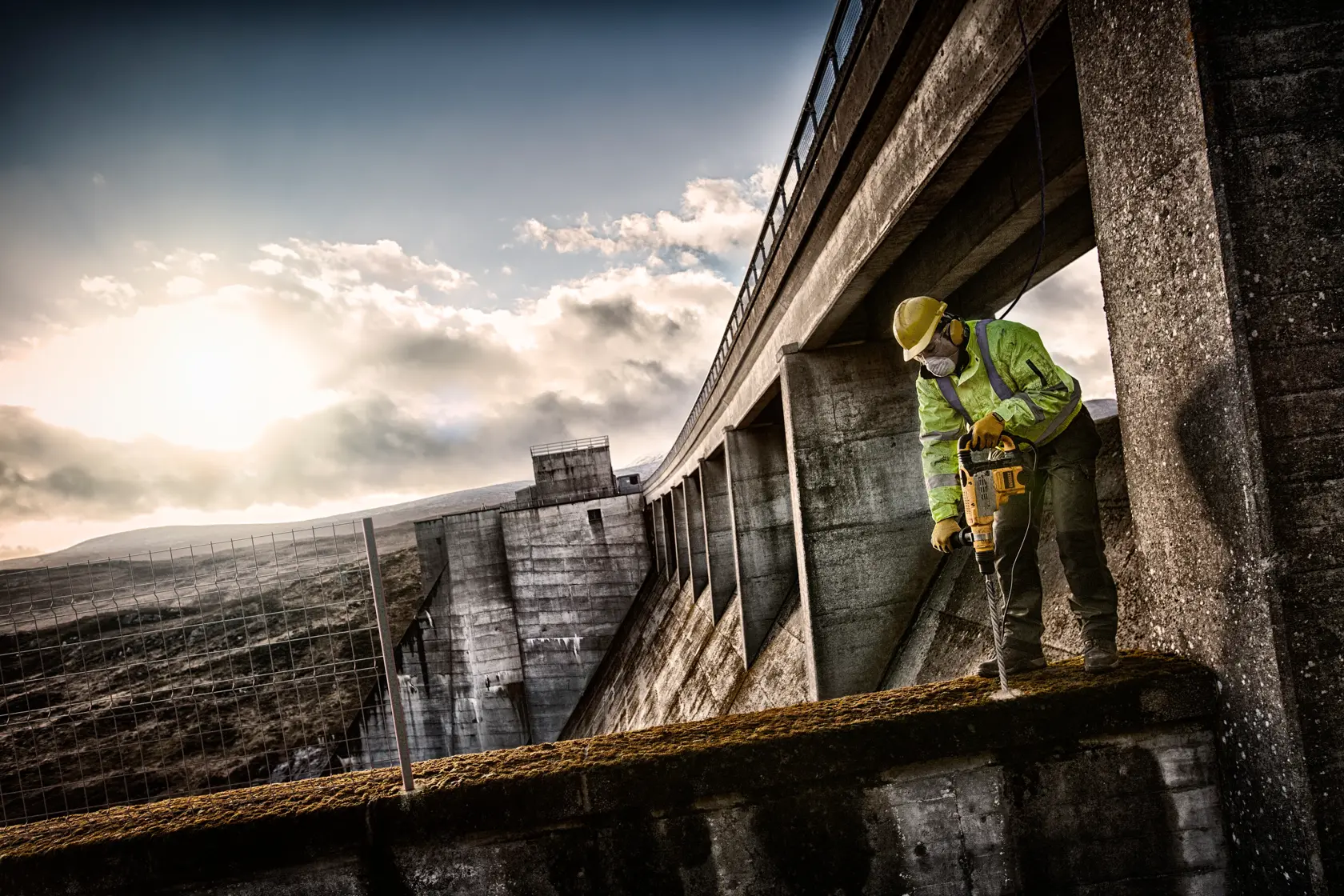 Arbeiter mit Schutzausrüstung bohrt Beton nahe einem großen Damm mit DEWALT Werkzeug.