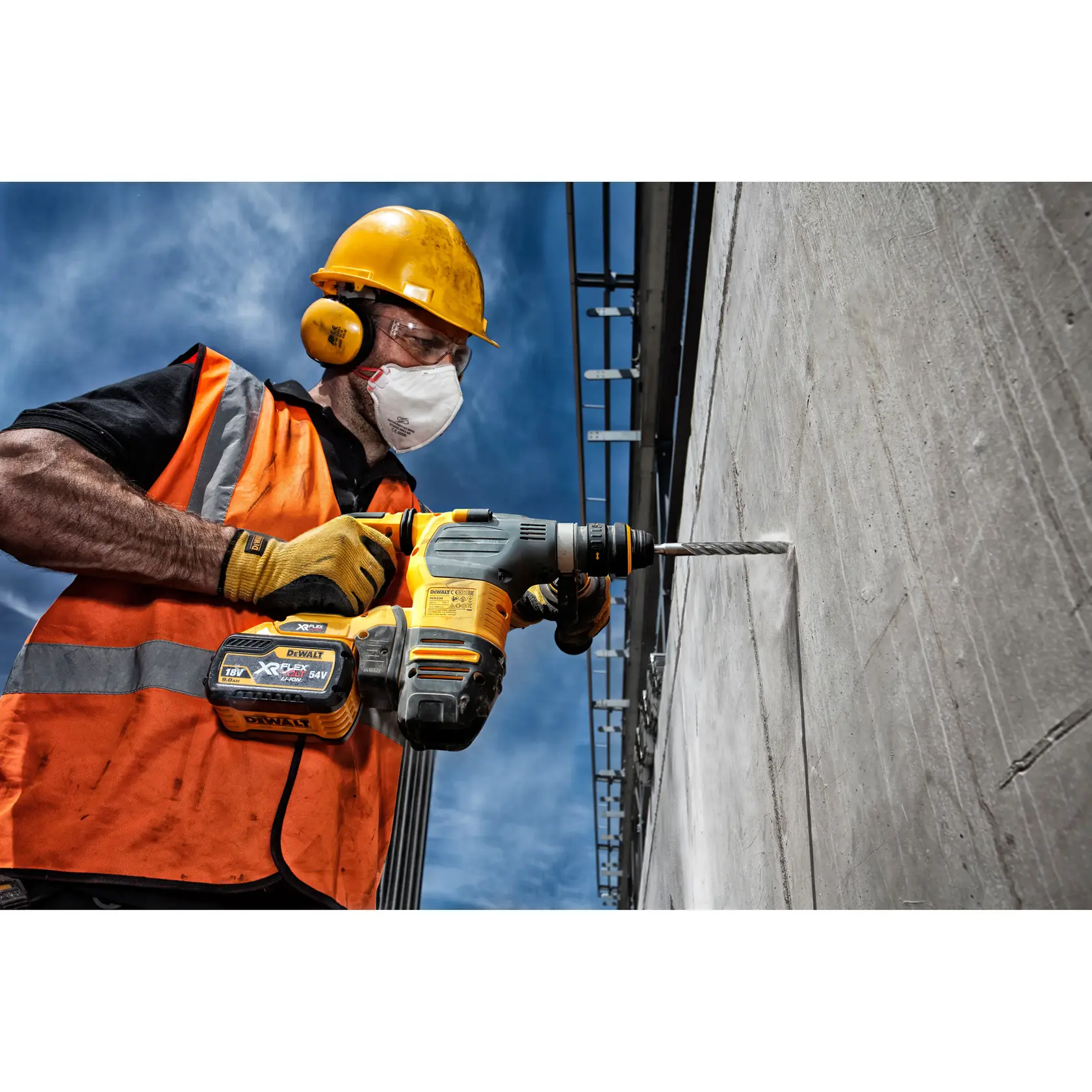 Worker wearing safety gear uses DEWALT cordless drill to bore into concrete wall outdoors under blue sky.